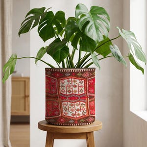 May include: A Monstera plant with large, green leaves in a cylindrical pot. The pot features a red, brown, and white patterned design, reminiscent of a rug. It rests on a small, round wooden stool. The pot's design is similar to a rug.