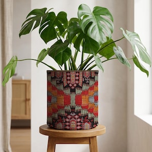 May include: A Monstera plant with large, green leaves in a cylindrical planter with a geometric pattern in red, orange, black, and grey. The plant pot is on a small, round wooden stool. The pot's pattern is reminiscent of a rug.