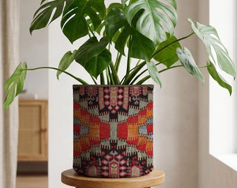 Puede incluir: Una gran planta de Monstera en maceta con hojas verdes vibrantes en una maceta cilíndrica. La maceta presenta un colorido patrón geométrico en rojo, naranja, negro y gris. La planta está sobre un taburete de madera redondo.