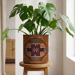 May include: A Monstera plant with large, green leaves in a brown woven planter. The planter has a geometric pattern in brown, orange, and burgundy. The plant pot is on a small, round wooden stool. The plant pot is approximately 30 cm in diameter.