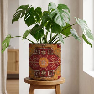 May include: A Monstera plant with large, green leaves in a patterned pot. The pot, in shades of red, yellow, and brown, sits on a small, round wooden stool. The pot's design resembles a rug. The plant is a decorative item.