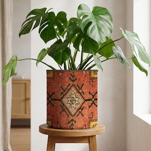 Op de afbeelding: Een Monstera-plant met grote, groene bladeren in een cilindrische plantenbak. De plantenbak heeft een rood, oranje en zwart geometrisch patroon, vergelijkbaar met een tapijt. De bloempot staat op een kleine, ronde houten kruk. De pot is ongeveer 25 cm hoog.