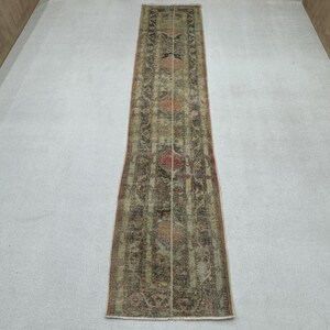 May include: A long, rectangular rug with an intricate, faded pattern in shades of green, brown, and red. The rug features a central design with floral and geometric motifs, and a distressed, vintage appearance. The rug is suitable for a hallway or entryway.
