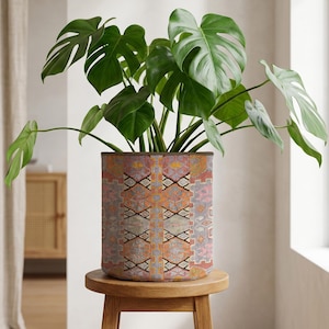 May include: A patterned plant pot containing a Monstera plant, set on a small wooden stool. The pot's design features geometric patterns in orange, pink, and grey. The plant has large, green leaves. The pot is cylindrical in shape.