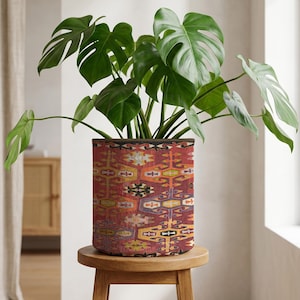May include: A Monstera plant with large, green leaves in a cylindrical planter. The planter has a patterned design in red, yellow, and blue. It sits on a small wooden stool. The planter's design is reminiscent of a rug.