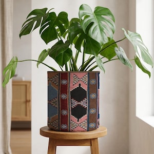 Könnte beinhalten: Eine große grüne Monstera-Pflanze in einem dekorativen zylindrischen Pflanzgefäß. Der Pflanztopf hat ein farbenfrohes geometrisches Muster in Rosa-, Blau-, Braun- und Schwarztönen. Der Pflanztopf steht auf einem kleinen, runden Holzschemel.
