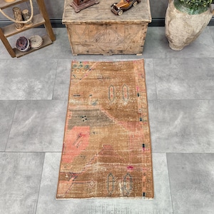 May include: A rectangular area rug featuring a distressed brown base with faded geometric patterns in muted pink, teal, and charcoal gray. The rug has a worn texture with visible signs of age, including frayed edges and uneven color distribution.