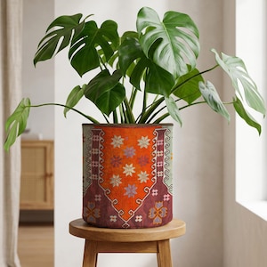 May include: A Monstera plant with large, green leaves in a cylindrical plant pot. The pot has a patterned design in orange, red, and floral motifs. The plant pot sits on a small, round wooden stool. The pot's diameter is approximately 30 cm.