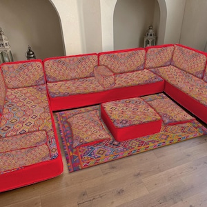 Peut inclure: Un canapé d'angle avec un tissu à motifs vibrants dans les tons de rouge, jaune et bleu. Le canapé a un cadre rouge et comprend un pouf et un tapis assortis, créant un ensemble de salon cohérent. Le design présente des motifs géométriques.