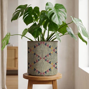 May include: A Monstera plant with large, green, split leaves is in a cylindrical planter. The planter has a textured, light-coloured background with a repeating geometric pattern of pink, blue, and yellow triangles. The planter sits on a small, round wooden stool.