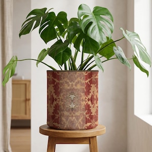 May include: A Monstera plant in a patterned pot, with large green leaves, sits on a small wooden stool. The pot's design features red, brown, and beige hues. The plant is indoors, near a window.