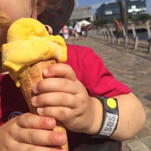 May include: A young child wearing a red shirt and a yellow hat holds a waffle cone filled with yellow ice cream. The child is wearing a blue and gray wristband with a yellow circle and the word "SOS" printed on it.