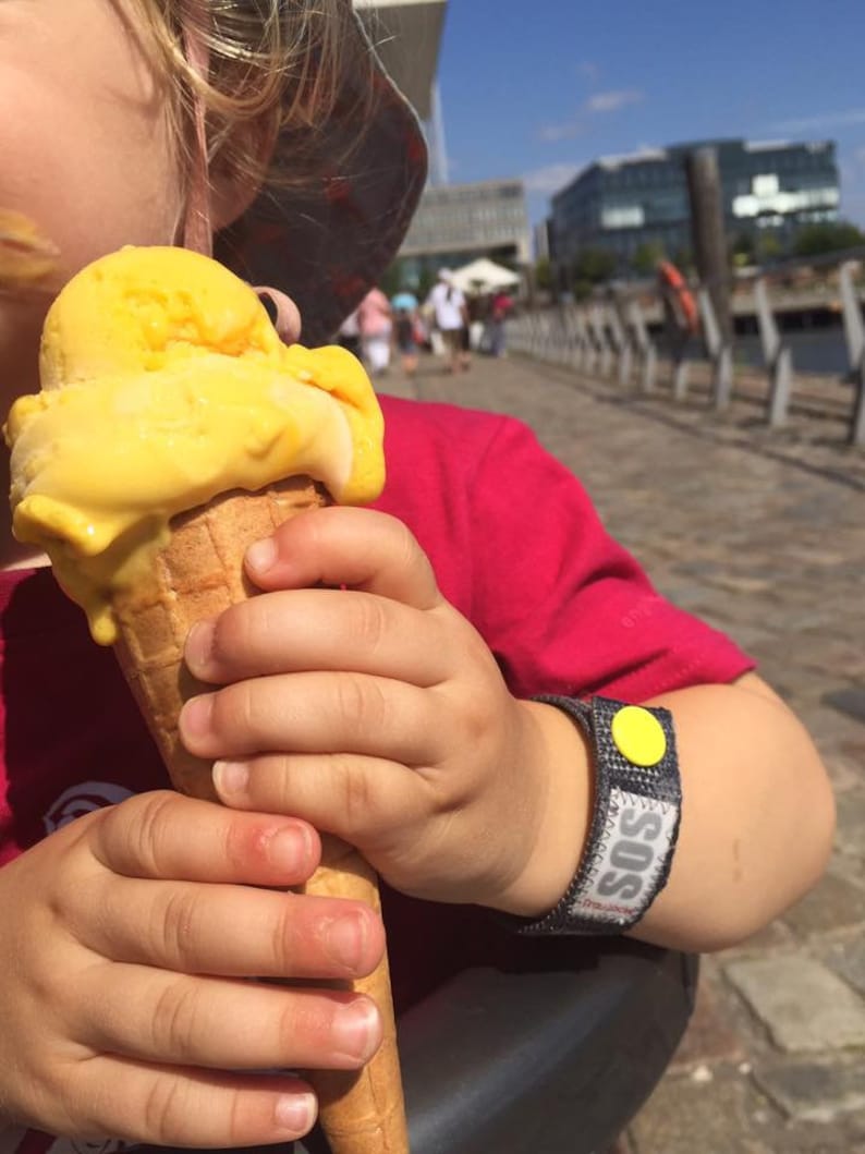 May include: A young child wearing a red shirt and a yellow hat holds a waffle cone filled with yellow ice cream. The child is wearing a blue and gray wristband with a yellow circle and the word "SOS" printed on it.