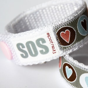 May include: White fabric bracelet with a pink button and the word "SOS" printed in gray. The bracelet also has a gray fabric band with brown circles and red and blue hearts.