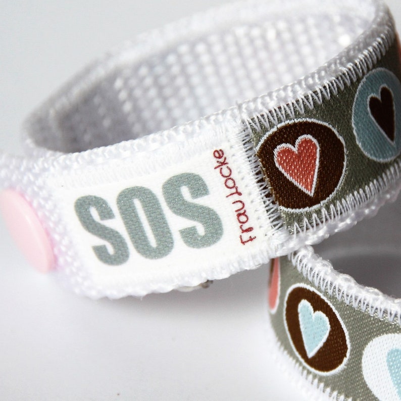May include: White fabric bracelet with a pink button and the word "SOS" printed in gray. The bracelet also has a gray fabric band with brown circles and red and blue hearts.