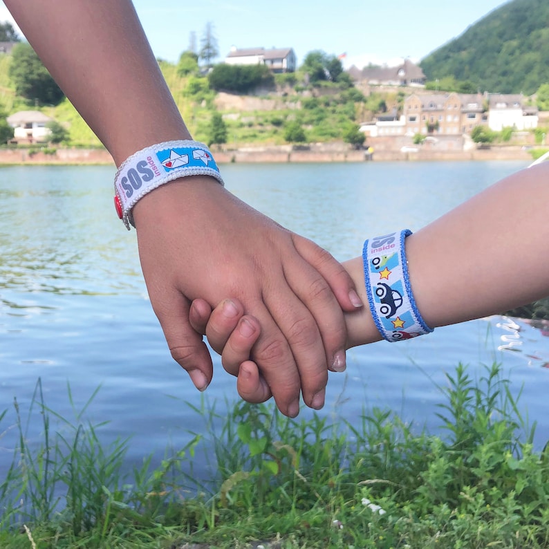 May include: Two children holding hands, each wearing a colorful wristband with the word "SOS" and a phone number printed on it. The wristbands are designed for safety and identification.