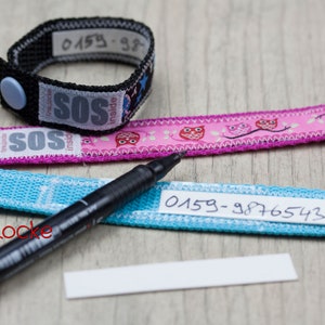 May include: Three fabric wristbands with a black marker pen and a blank white label. The wristbands are blue, pink, and black. The blue and pink wristbands have a white label with black writing that says "0159-9876543". The black wristband has a white label with black writing that says "0159-98". The pink wristband has a pink button and the blue wristband has a blue button. The black wristband has a blue button. The pink wristband has a pink owl design. The wristbands are for identification.