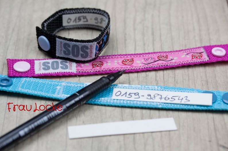 May include: Three fabric wristbands with a black marker pen and a blank white label. The wristbands are blue, pink, and black. The blue and pink wristbands have a white label with black writing that says "0159-9876543". The black wristband has a white label with black writing that says "0159-98". The pink wristband has a pink button and the blue wristband has a blue button. The black wristband has a blue button. The pink wristband has a pink owl design. The wristbands are for identification.