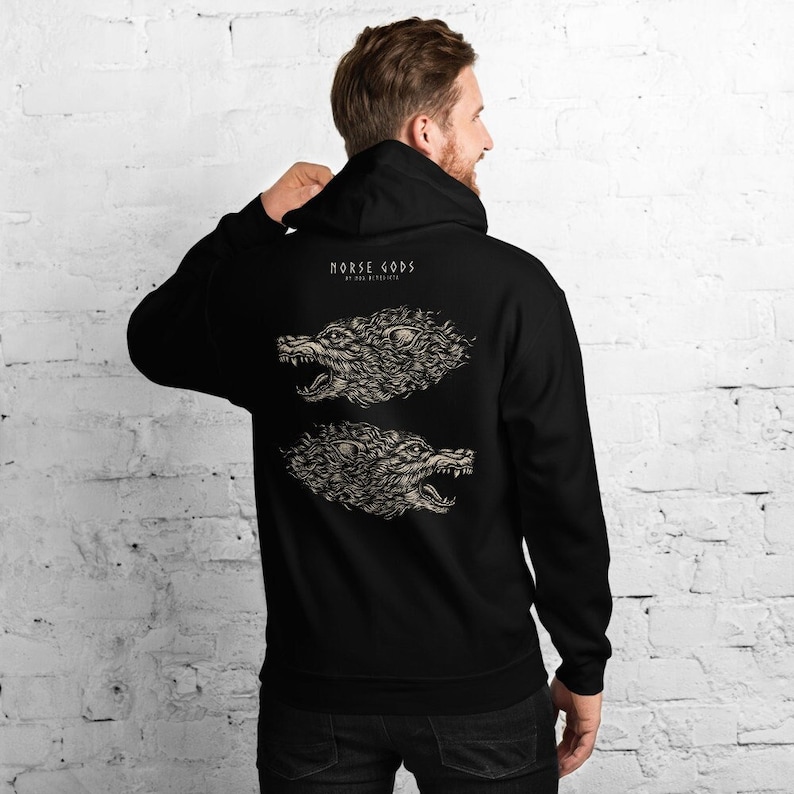 Puede incluir: Una sudadera con capucha negra que presenta un gr&aacute;fico en la espalda con el texto "NORSE GODS" sobre dos ilustraciones detalladas y reflejadas de cabezas de lobo gru&ntilde;endo en color beige claro, con un estilo art&iacute;stico texturizado.