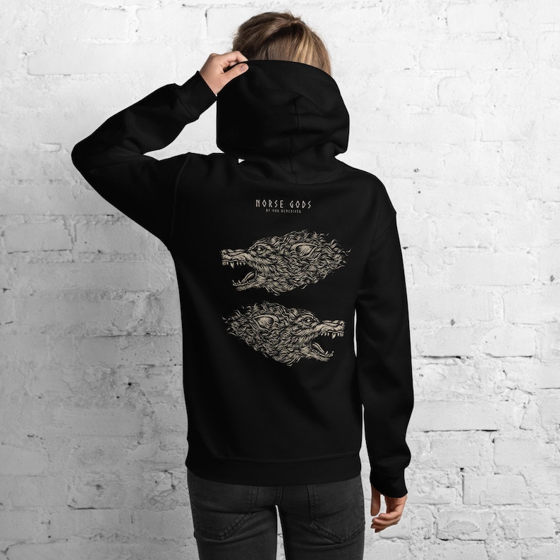 Puede incluir: Una sudadera con capucha negra que presenta un estampado gr&aacute;fico en la parte posterior. El dise&ntilde;o incluye el texto "NORSE GODS" sobre dos ilustraciones detalladas y reflejadas de cabezas de lobo gru&ntilde;endo en color beige claro sobre la tela.