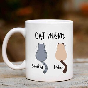 May include: White ceramic mug with the text "CAT MOM" in black lettering. Two cartoon cats are shown sitting back-to-back with their names "Smokey" and "Timber" written below them.