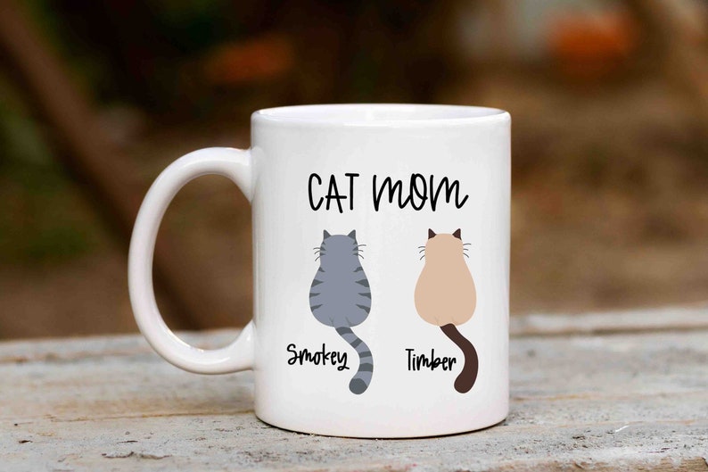 May include: White ceramic mug with the text "CAT MOM" in black lettering. Two cartoon cats are shown sitting back-to-back with their names "Smokey" and "Timber" written below them.