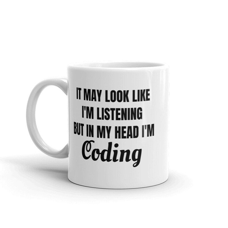 May include: White ceramic mug with a curved handle. The mug features black text that reads: "IT MAY LOOK LIKE I'M LISTENING BUT IN MY HEAD I'M Coding". The mug is designed for programmers and coders.