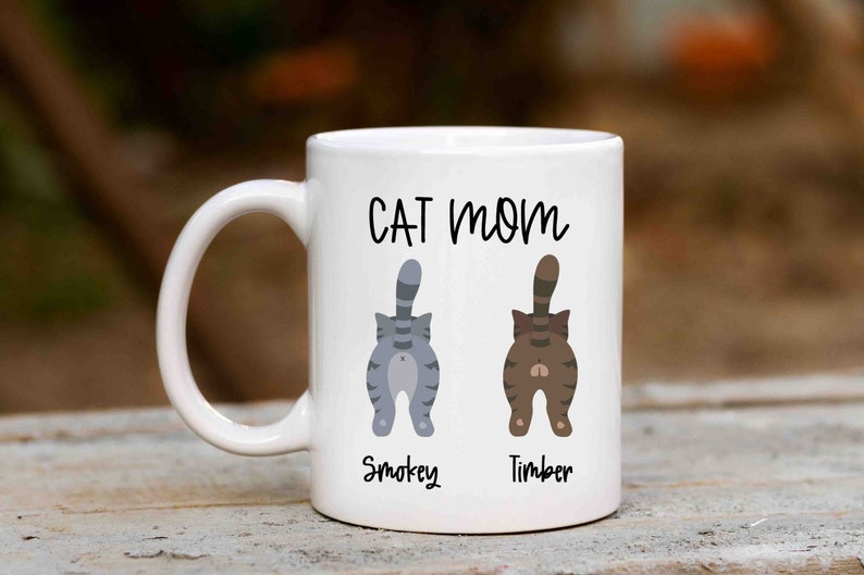 May include: White ceramic mug with the text "CAT MOM" printed in black. Two cartoon cats are shown with their backs to the viewer, one gray and one brown, with their names "Smokey" and "Timber" printed below them.