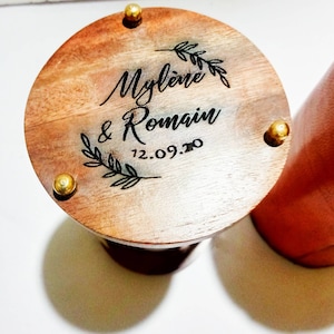 May include: A wooden, round container lid with the names "Mylène & Romain" and the date "12.09.20" engraved. The lid is adorned with a leaf design and secured with three gold-colored studs. The container's side is a reddish-brown color.