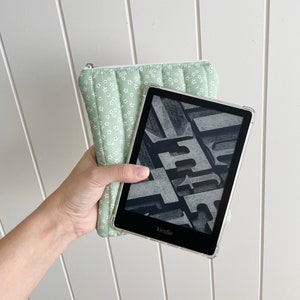 May include: A green floral patterned case with a black Kindle e-reader inside. The Kindle screen displays a black and white image of text.