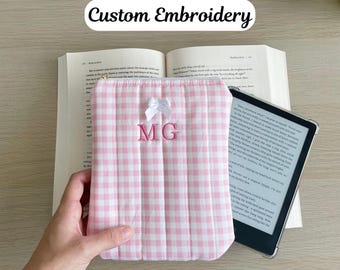 以下が含まれることがあります： ピンクと白のギンガムチェック柄のブックカバー。白いリボンと刺繍された「MG」のイニシャルが付いています。カバーはキルティング加工が施され、柔らかくパッドが入った外観です。「Custom Embroidery」の文字が上部にあります。
