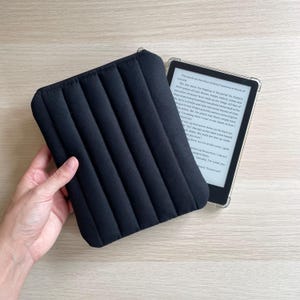 May include: Black padded e-reader case with a zip closure and a quilted design. An e-reader in a clear case is next to the case. The case has vertical lines.