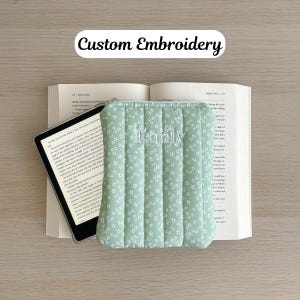 May include: A green quilted e-reader case with a white floral pattern and the name "Emily" embroidered on the front.