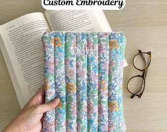 May include: A floral-patterned book sleeve with the text "Custom Embroidery" and the embroidered name "Olivia". The sleeve is filled with a book and is next to a pair of eyeglasses.