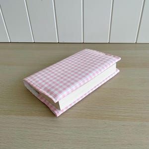 May include: A pink and white gingham book cover. The cover is made of fabric and has a white book inside.