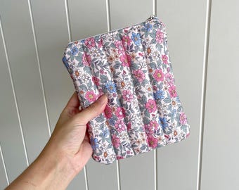 May include: A floral-patterned quilted pouch with a zipper closure. The pouch features a white background with pink, blue, and white flowers. The pouch is held against a light gray wooden background.