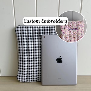 May include: A silver iPad sits beside a black and white gingham tablet sleeve. The sleeve is open, showing the inside. A smaller, circular image shows a floral tablet case with the name "Jessica" embroidered. The text "Custom Embroidery" is above the iPad.