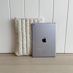 May include: A white quilted tablet case with a floral pattern and a silver iPad with the Apple logo on the back. The iPad is sitting on a wooden surface.