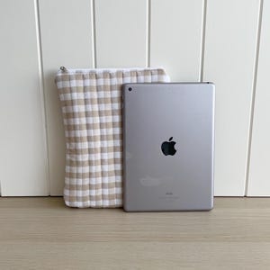 May include: A beige and white gingham tablet case with a zipper closure. The case is next to a silver iPad with the Apple logo.