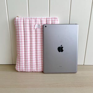 May include: A pink and white gingham iPad case with a white bow. The case is next to a silver iPad with the Apple logo.