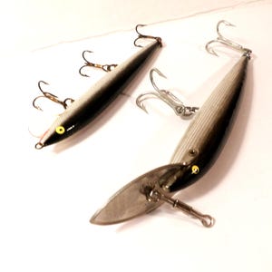 May include: Two fishing lures with a black and silver color scheme. Each lure has multiple treble hooks and a small, yellow eye. One lure has a silver, paddle-like attachment. The lures are designed for catching fish.