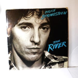 May include: Vintage vinyl record album cover featuring a black and white portrait of Bruce Springsteen. The album title "The River" is in blue text, with Springsteen's name above. The cover has a worn appearance.