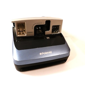 May include: A vintage Polaroid One600 instant camera. The camera has a blue and black body with a silver top. The Polaroid logo is visible on the front. The camera has a flash and a lens. The focus range is 3ft - ∞.