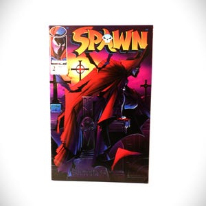 May include: A Spawn comic book issue number 2 with a vibrant cover illustration. The artwork depicts a cloaked figure in a graveyard at sunset, featuring a large red cape, dark silhouettes of crosses, and the bold yellow Spawn logo with a skull icon.