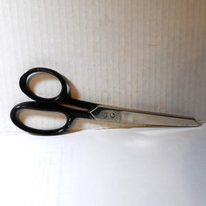 May include: A pair of scissors with black handles and silver blades. The scissors are lying on a white surface with a cardboard background. The blades are sharp and the handles have finger holes.