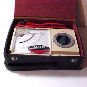 May include: An open, vintage electrical testing device in a black leatherette case with a burgundy interior. The device features a meter with a white face, a dial, and red wires. The text "MGC" is visible on the meter.