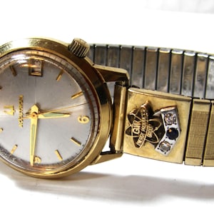 May include: A vintage gold-toned wristwatch with a silver face and gold hands. The watch has a stretch band and a decorative clasp with the text "GW Energy Products Group". The watch is a classic timepiece.
