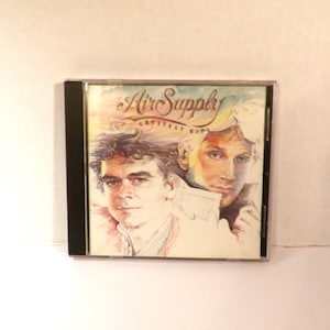 May include: An Air Supply Greatest Hits music CD in a standard clear plastic jewel case. The cover art features a watercolor-style illustration of two individuals with light hair, set against a soft, pastel-toned background with cursive text.