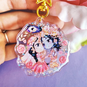 May include: A clear resin keychain with a gold star charm and a colorful illustration of two figures holding hands. The figures are surrounded by flowers and glitter.
