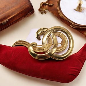 May include: A gold-toned metal brooch with an interlocking design, resting on a red velvet pillow. The brooch features a circular and curved shape with intricate detailing. Other antique items are in the background.